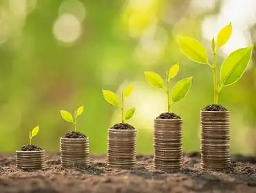 Five stacks of coins arranged in ascending order, each topped with a small plant sprouting leaves, symbolizing growth and financial prosperity. The background is blurred with bright green and yellow hues, suggesting a natural, sunny environment.