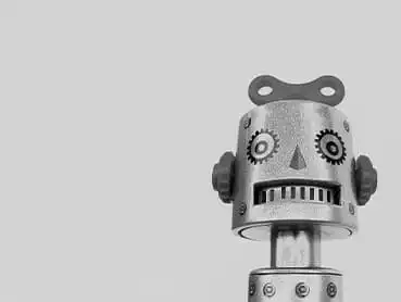 A monochrome image of a vintage tin robot with a wind-up key on top of its head. The robot has a cylindrical body, round eyes with gear-like details, a triangular nose, and a smiling mouth with rectangular teeth. It stands against a plain background.